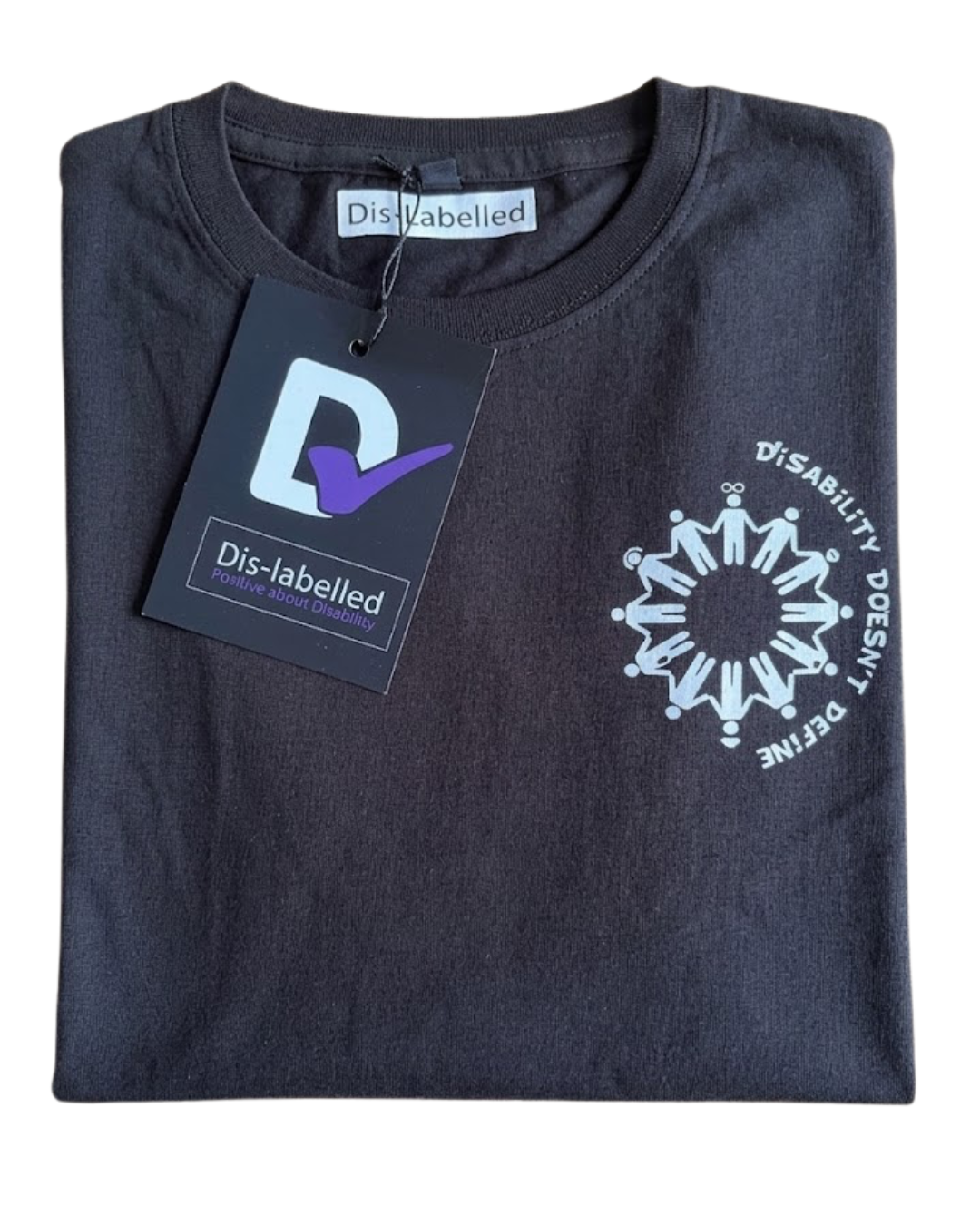 A neatly folded black Dis-Labelled charity t-shirt displayed flat on a light wooden background. The shirt features a white circular logo on the left chest area depicting a ring of stick figures holding hands, surrounded by the text "DiSABILITY DOESN'T DEFINE" in all caps. A black cardboard clothing tag with a prominent white 'D' and a purple checkmark hangs from the collar, partially covering the upper left side of the shirt.